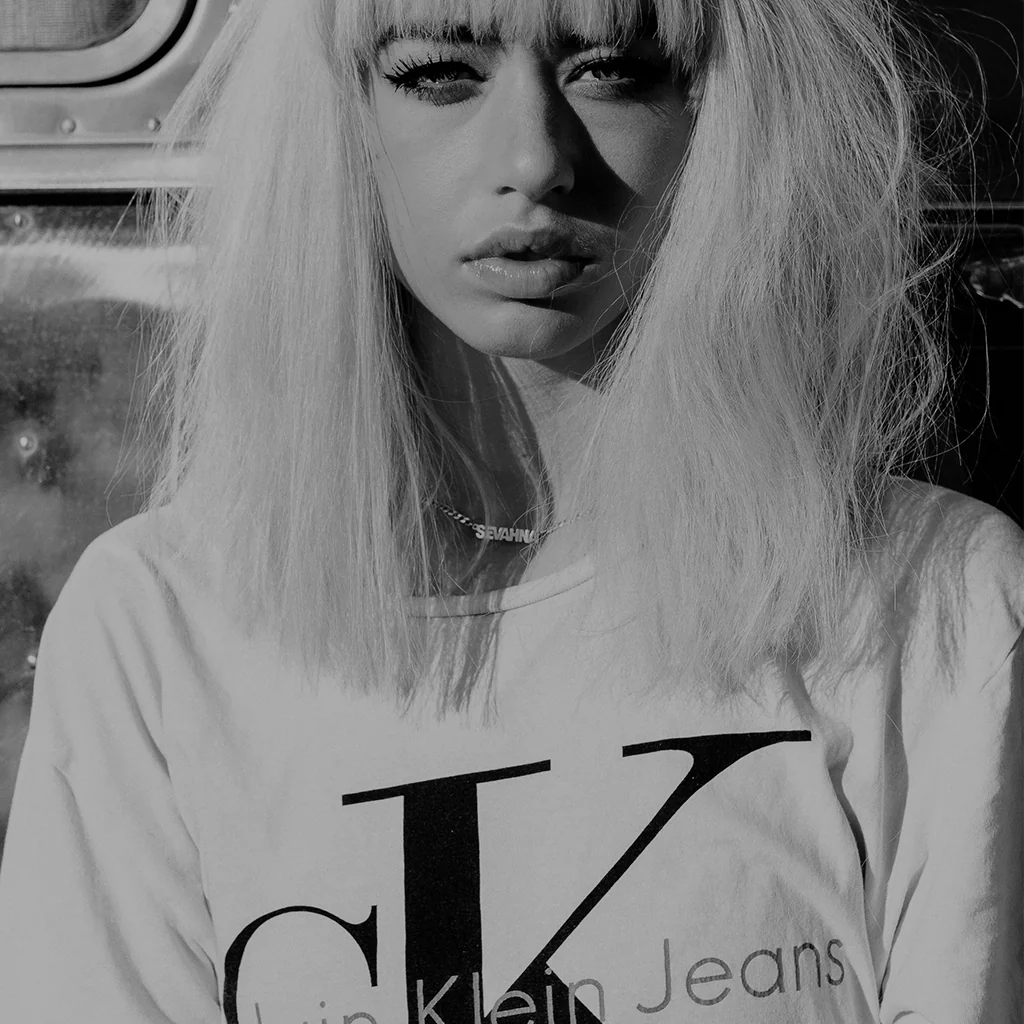 Woman wearing a Calvin Klein Jeans t-shirt with straight blonde hair standing in natural light against a metallic background