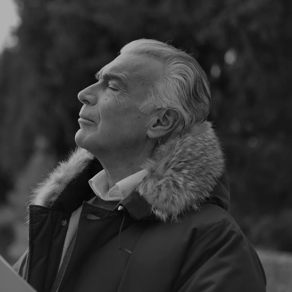Man wearing a fur lined winter jacket and collared shirt with eyes closed, standing outdoors against a blurred background of trees
